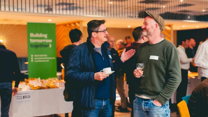 Two middle ages men chatting. Behind them is a stall with a green banner