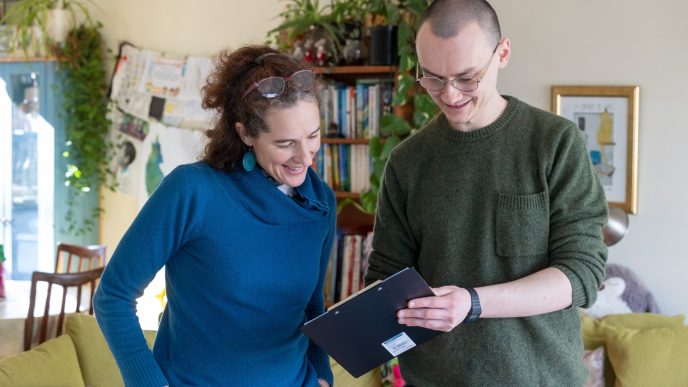 Photo of smiling, bald man in green jumper holding a clipboard and showing it to smiling, woman with warm, brown hair and glasses on head and blue top in a living room