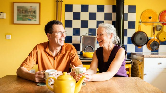 Smiling couple holding cups of tea in their hands, with a yellow tea pot in front of them. Behind them is an aga and some kitchen pots, with yellow walls.