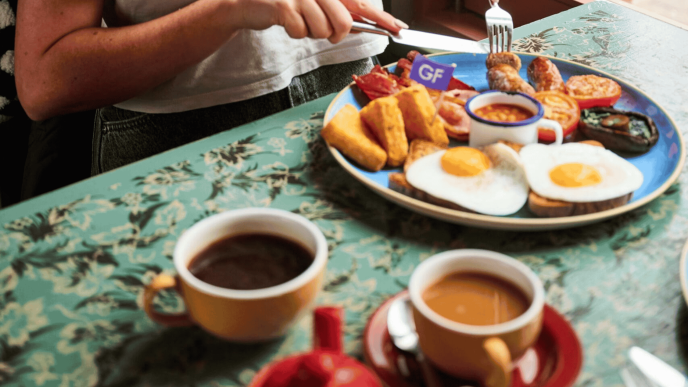 Photo of cooked breakfast (eggs, toast, hash browns, sausages. bacon, beans, tomato, mushroom) on a table with tea and coffee. Hands with knife and fork cutting into breakfast.